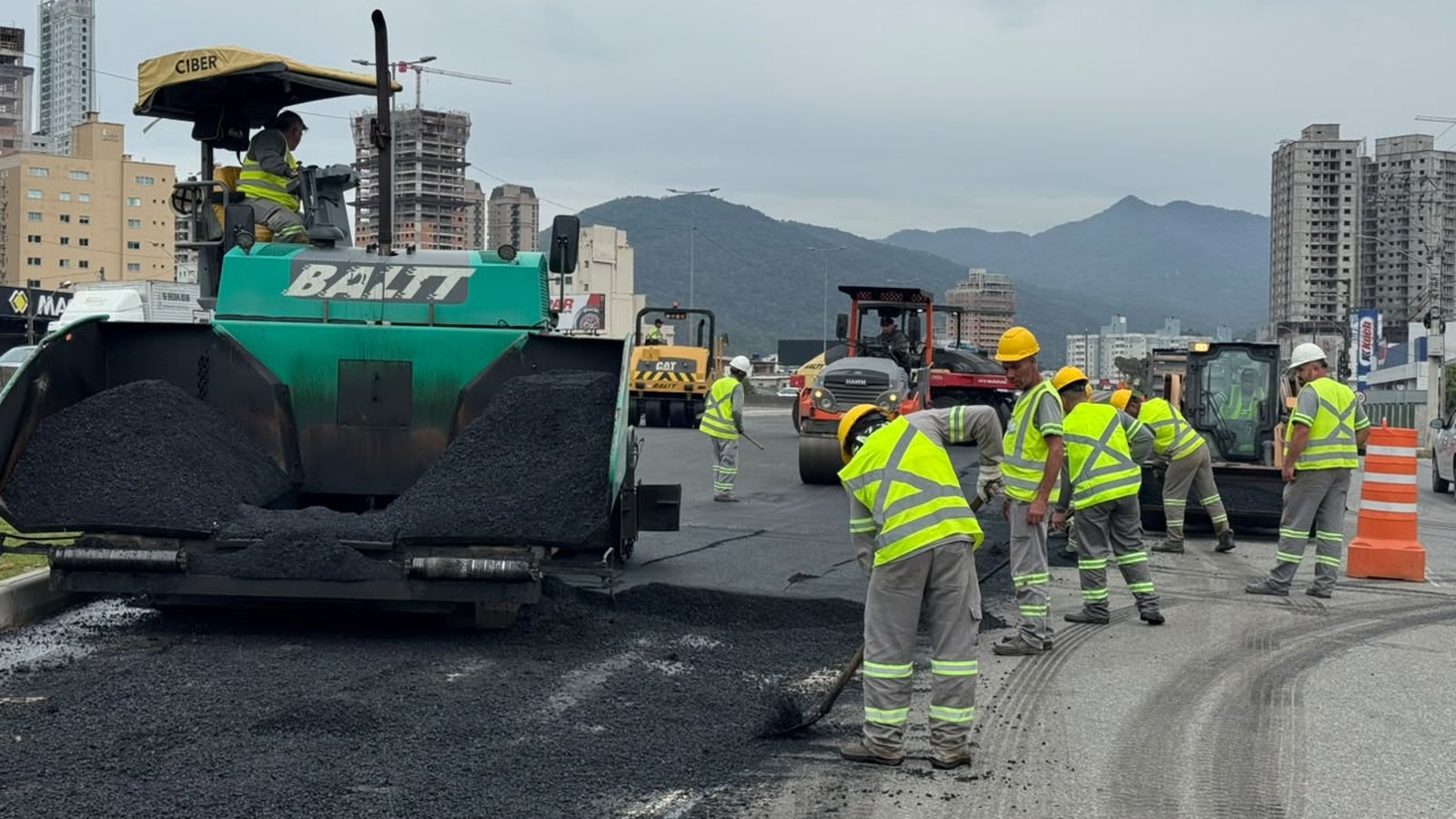 Pavimentadora verde da empreiteira BALTT espalhando asfalto quente na alça da BR-101 em Itapema, com seis operários de colete refletivo e capacete trabalhando ao lado, rolo compactador e retroescavadeira ao fundo, prédios da cidade e morro na linha do horizonte.
