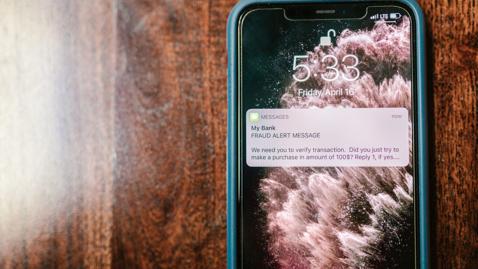 Celular apoiado sobre uma mesa de madeira escura, com a tela bloqueada mostrando uma notificação de mensagem que se apresenta como aviso de fraude do banco e pede confirmação de uma compra.