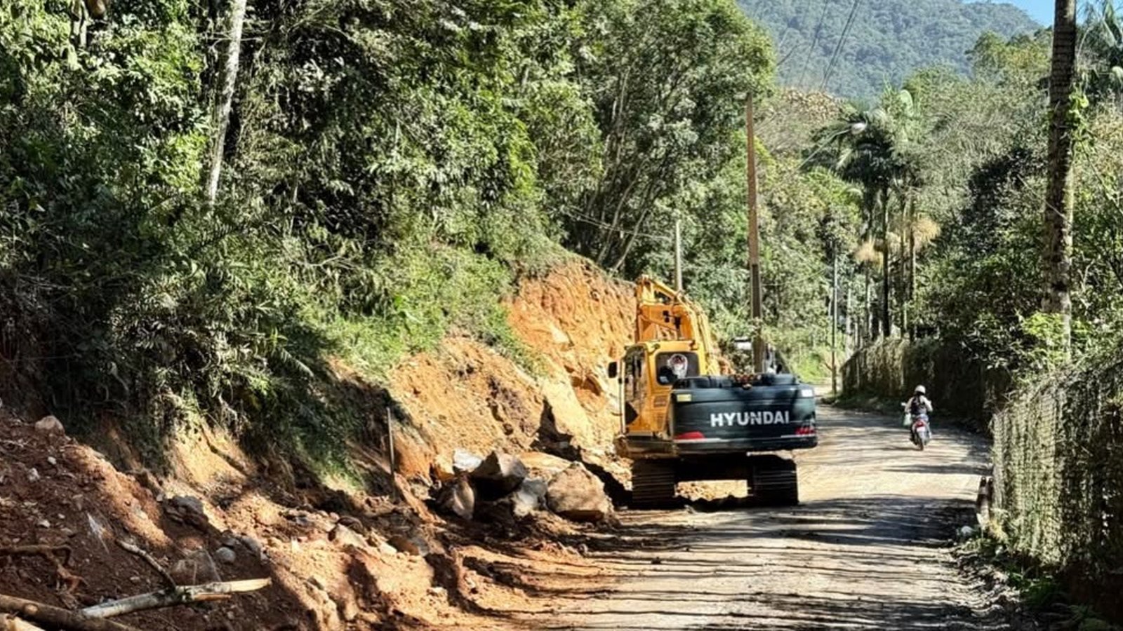 Escavadeira amarela da marca Hyundai cortando o barranco de terra vermelha na margem de uma estrada rural estreita e sem asfalto no Alto Areal, em Itapema, com blocos de rocha soltos no chão, mata fechada dos dois lados, poste de energia e um motociclista passando ao fundo.