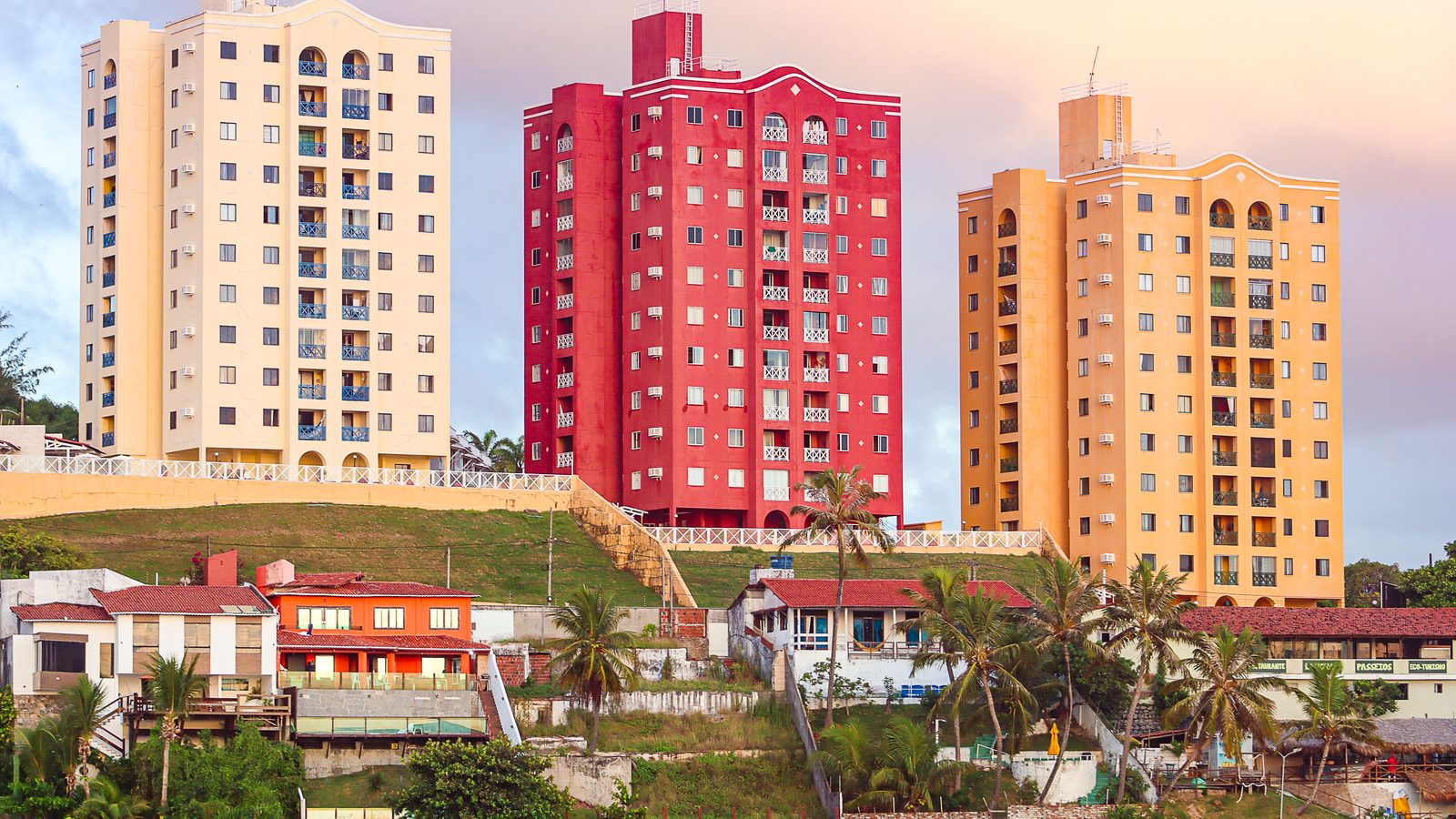 Três prédios residenciais de mais de dez andares, um bege, um vermelho e um amarelo, alinhados no alto de um morro à beira-mar, com casas baixas, coqueiros e um restaurante na encosta abaixo deles.