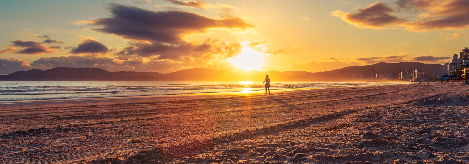 Nascer do sol sobre uma praia de Itapema: o sol baixo tinge o céu e a areia molhada de laranja, uma pessoa aparece em silhueta na beira da água e a linha de prédios surge ao longe, à direita.