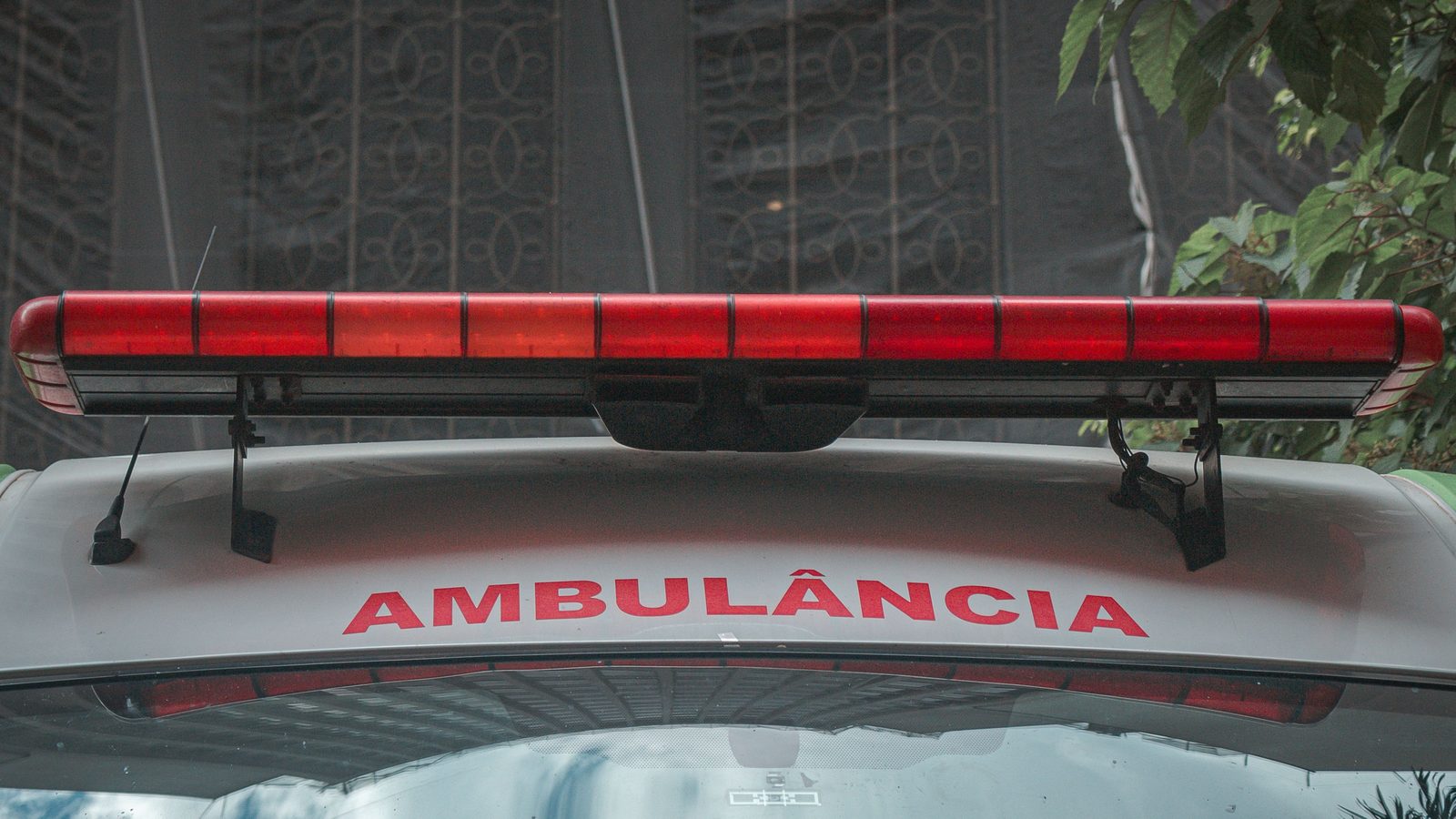 Vista frontal do teto de uma ambulância branca, com a barra de sirene vermelha atravessando a imagem e a palavra AMBULÂNCIA escrita em letras vermelhas sobre o capô.