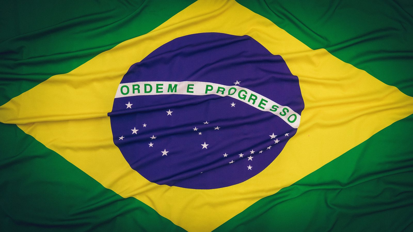 Bandeira do Brasil estendida e amassada, ocupando todo o quadro, com o losango amarelo, o círculo azul e a faixa branca escrita Ordem e Progresso.