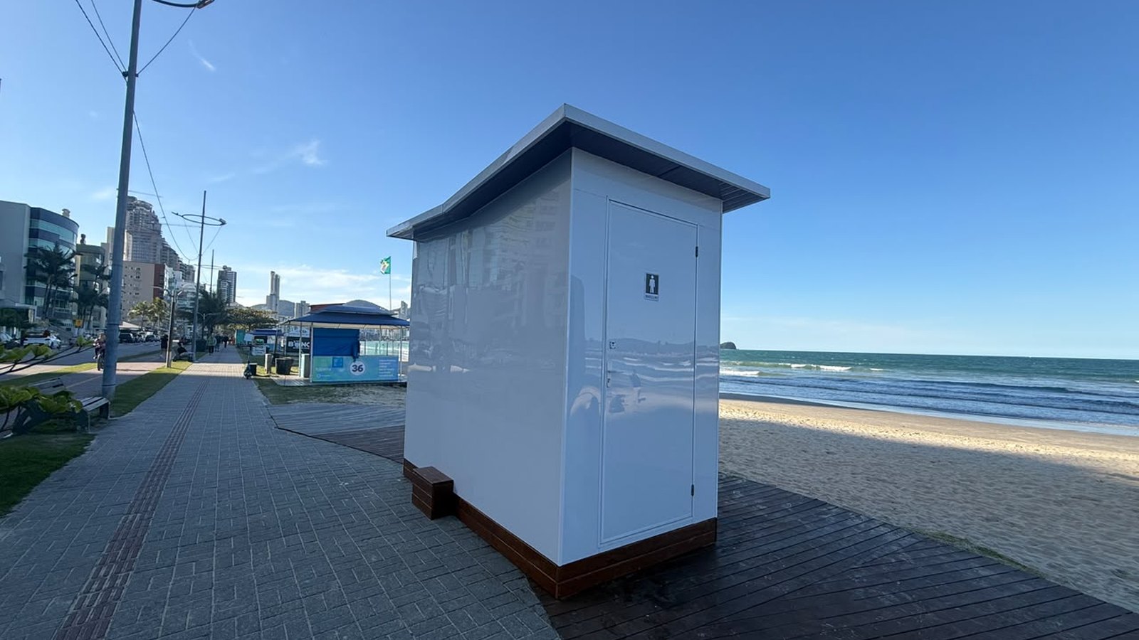 Módulo sanitário branco de porta única já instalado sobre o deque de madeira do calçadão da orla de Itapema, com a faixa de areia e o mar à direita e a linha de prédios da avenida à esquerda.