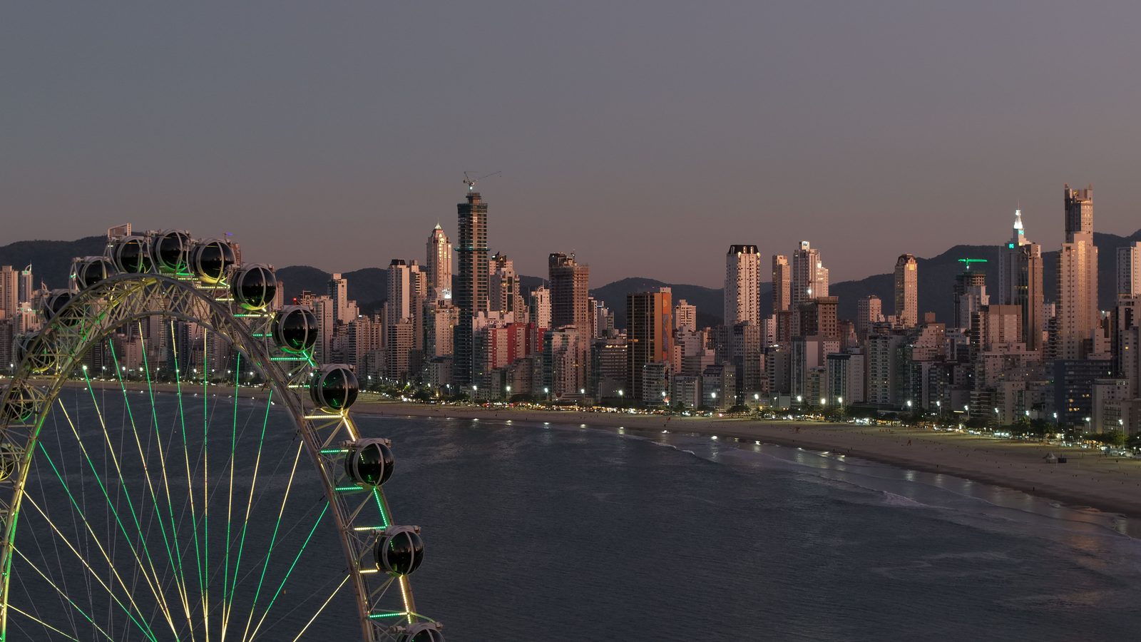 Balneário Camboriú ao anoitecer vista do alto: a faixa de prédios altos acompanha a curva da praia, com uma roda-gigante iluminada em primeiro plano e o mar escuro à direita.