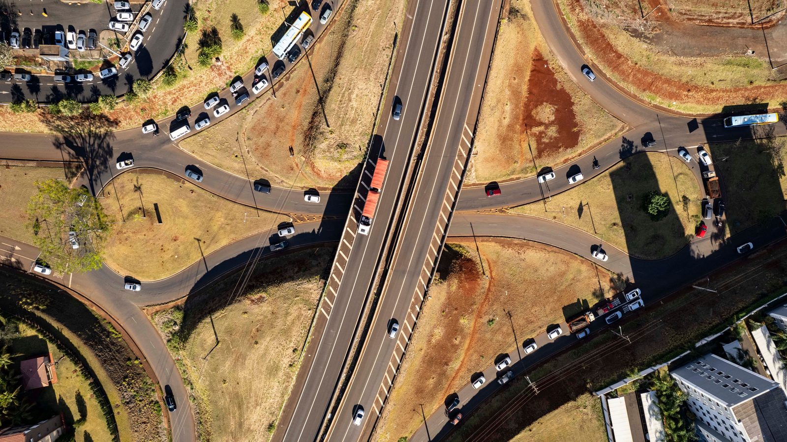 Vista aérea de um trevo de rodovia, com a pista principal cortando o quadro na diagonal e as vias marginais e alças de acesso cheias de carros parados dos dois lados.