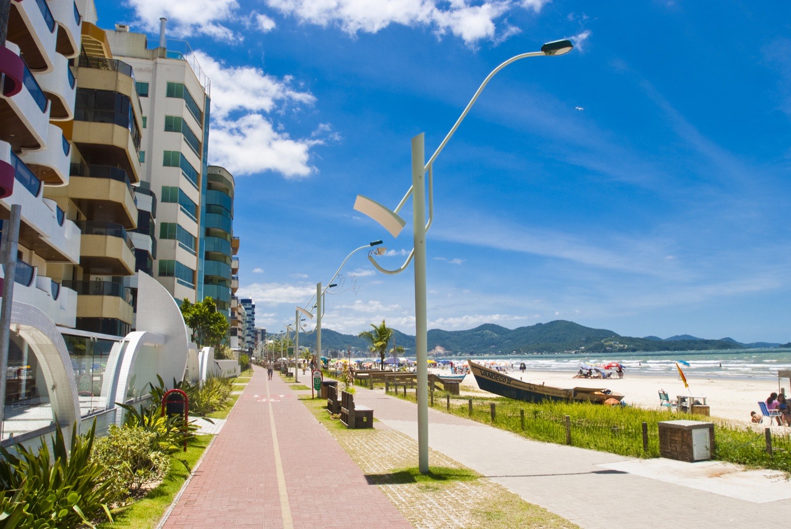 Calçadão da Meia Praia em dia de sol, com ciclovia de piso vermelho, postes de iluminação, prédios residenciais à esquerda e a faixa de areia e o mar à direita.