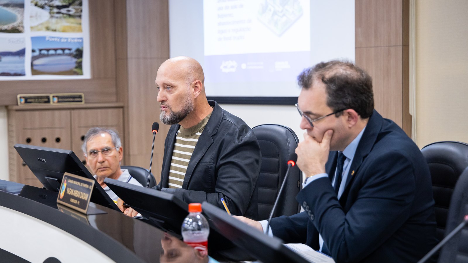 Mesa da audiência pública no plenário da Câmara de Itapema, com o vereador que preside a comissão falando ao microfone e, ao fundo, uma tela projetando slides sobre legislação urbanística.