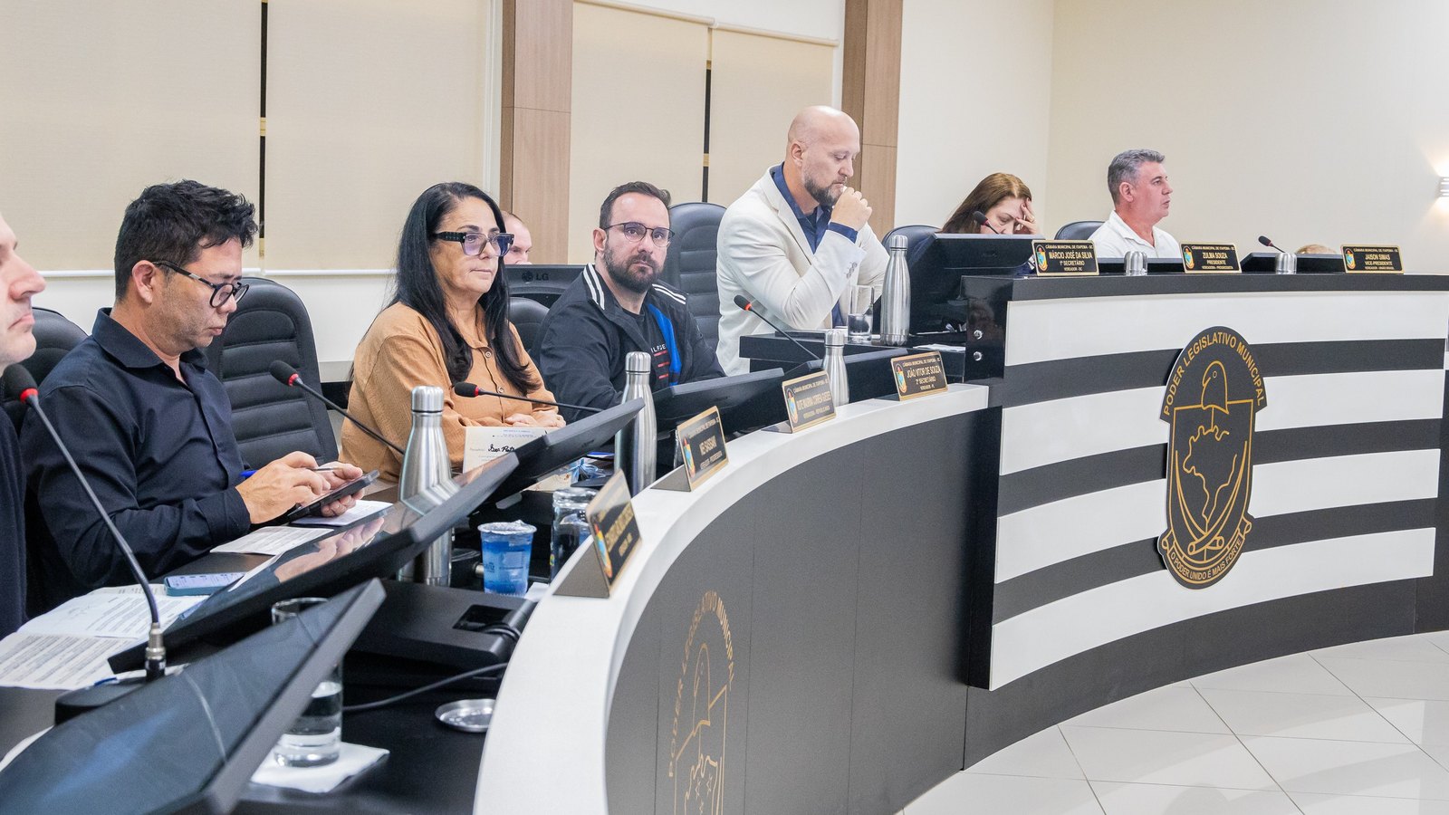 Vereadores sentados na bancada em curva do plenário da Câmara de Itapema durante sessão, com microfones, placas de identificação e o brasão do Poder Legislativo Municipal na lateral.