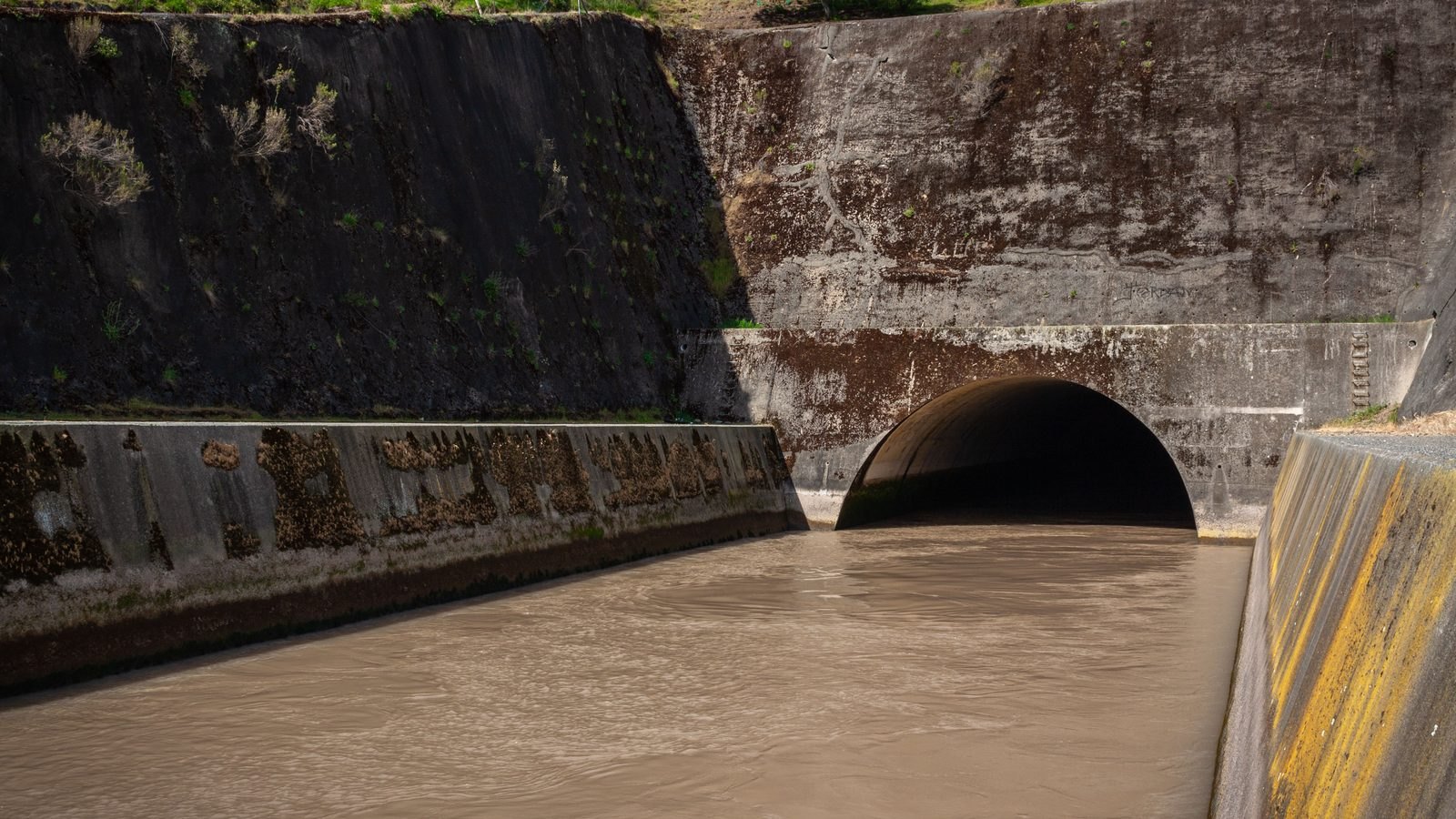 Canal de concreto com água barrenta correndo para dentro de uma galeria em arco, com as paredes laterais marcadas pelo nível antigo da água e mato nascendo no talude de cima.