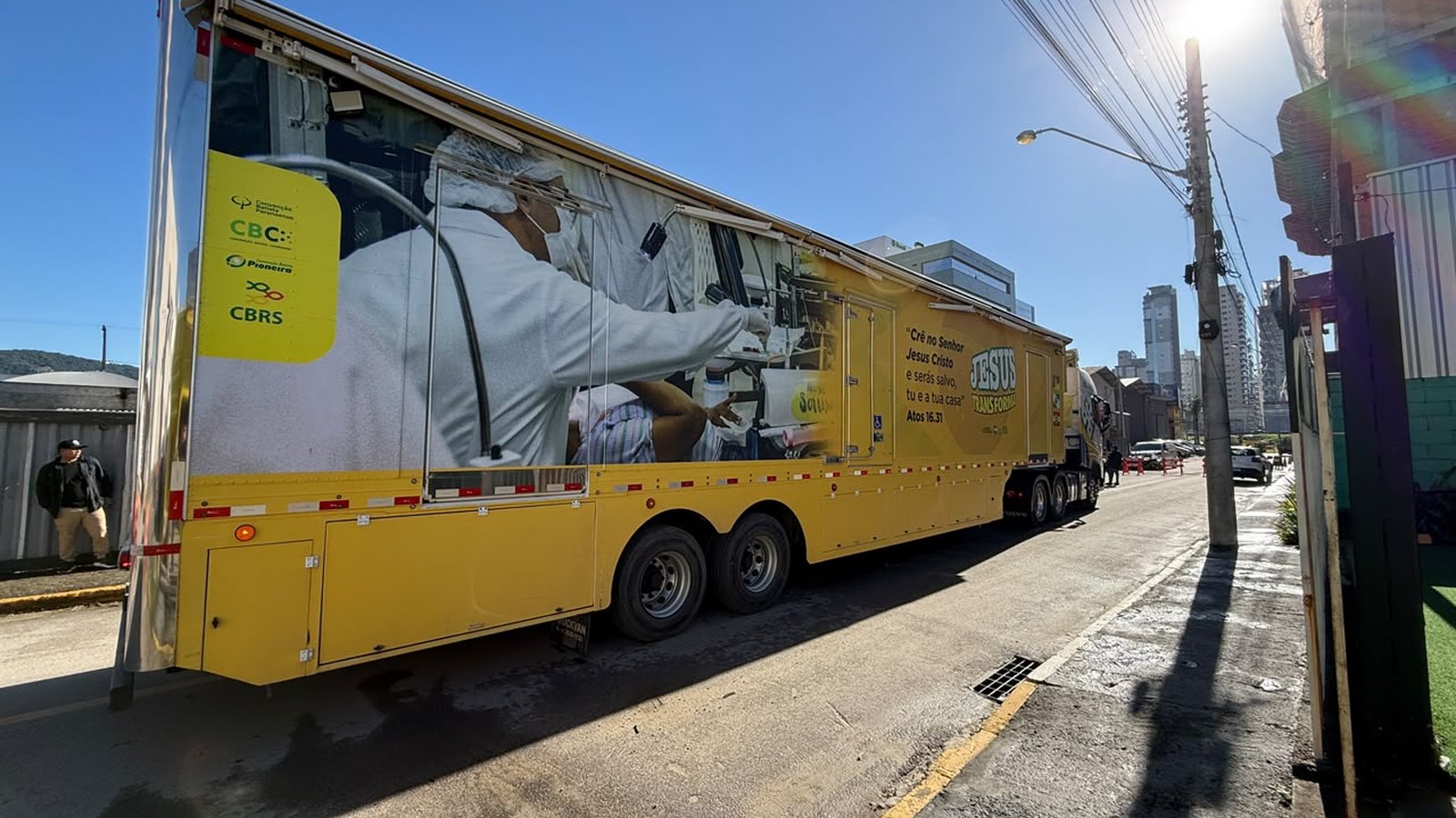 Carreta amarela de atendimento de saúde estacionada em rua de Itapema, com a lateral estampada por uma foto de profissional em atendimento, e prédios da cidade ao fundo.