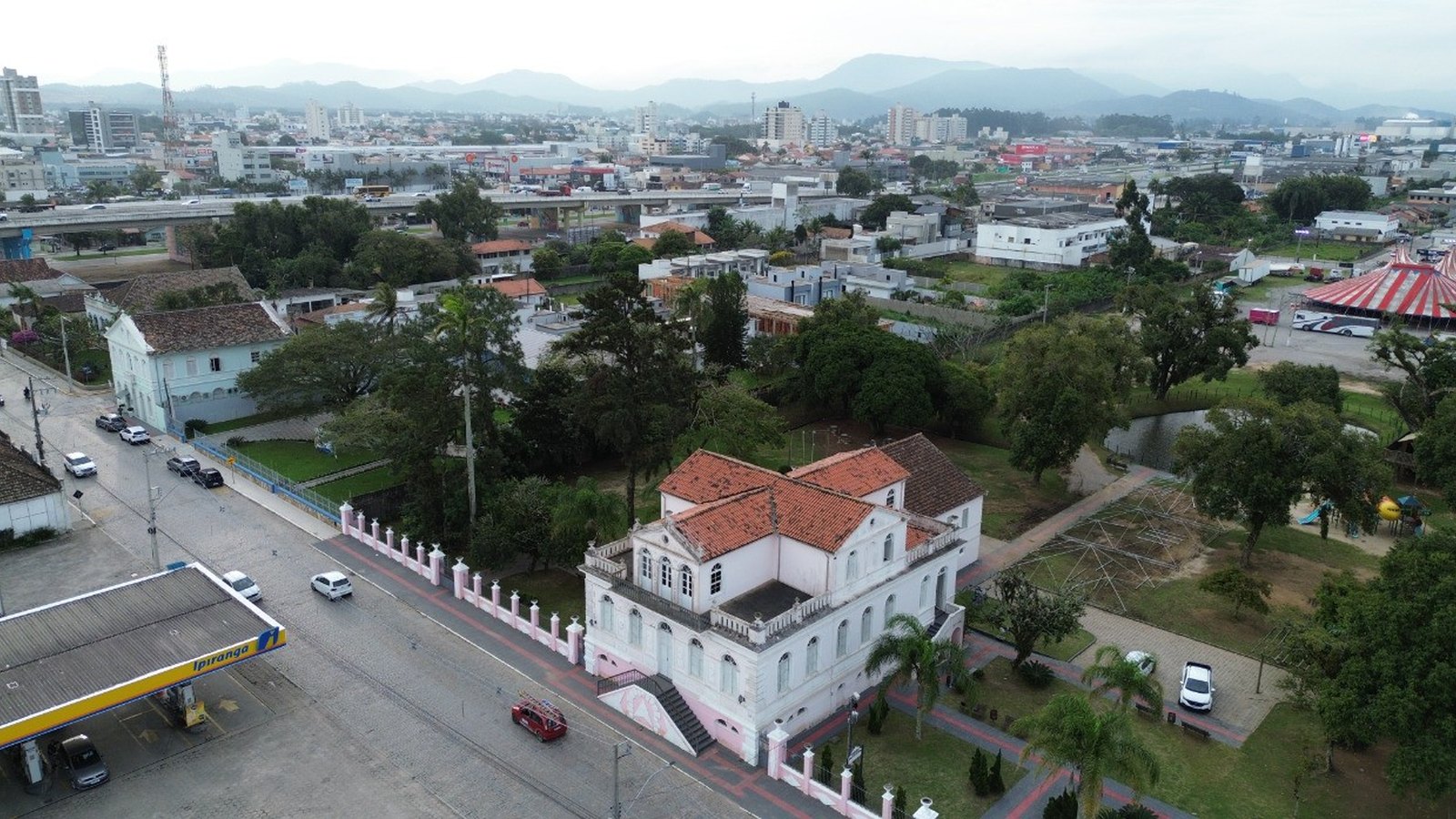 Vista aérea do Casarão Gallotti, sobrado histórico de paredes rosa claro e telhado de barro, cercado por um muro de pilaretes rosa e branco, com avenida e posto de combustível de um lado e parque arborizado do outro, e a cidade de Tijucas ao fundo.