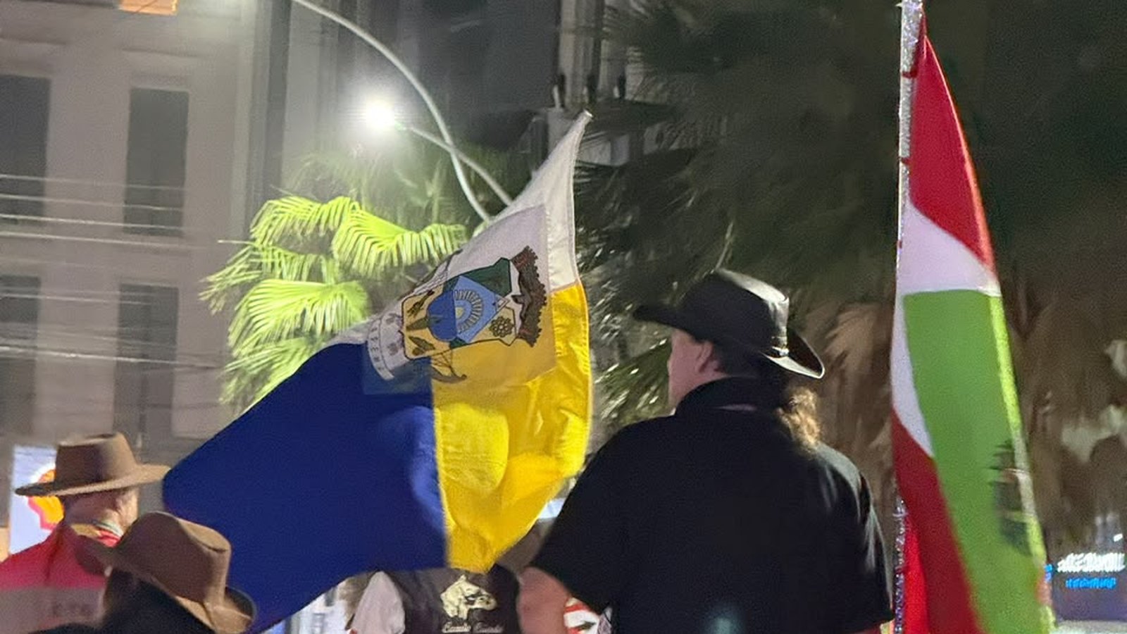Cavalgada à noite numa avenida de Itapema. Cavaleiros de chapéu, vistos de costas, seguem lado a lado carregando duas bandeiras grandes, uma azul e amarela com brasão e outra em faixas verde, branca e vermelha, com palmeiras e prédios iluminados ao fundo.