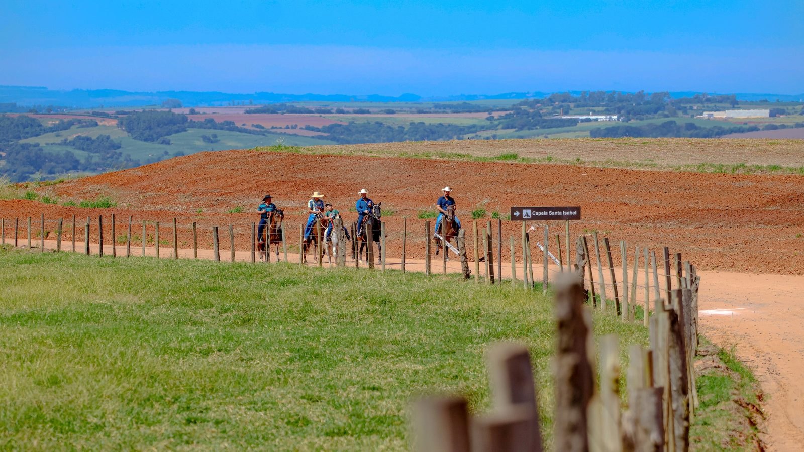 Quatro cavaleiros de chapéu montados em fila numa estrada de terra, vistos de longe, com cerca de mourões de madeira em primeiro plano, campo verde de um lado, terra vermelha lavrada do outro e morros baixos no horizonte sob céu azul.