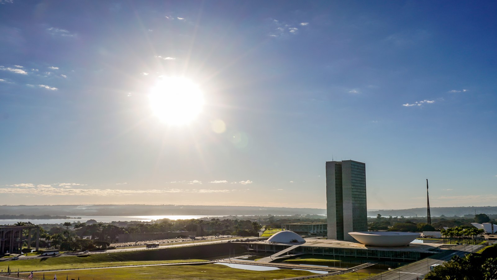 Vista aérea da Esplanada dos Ministérios em Brasília ao amanhecer, com o sol baixo no céu azul, o edifício alto do Congresso Nacional e as duas cúpulas branca do Senado e da Câmara ao lado dos espelhos d'água.