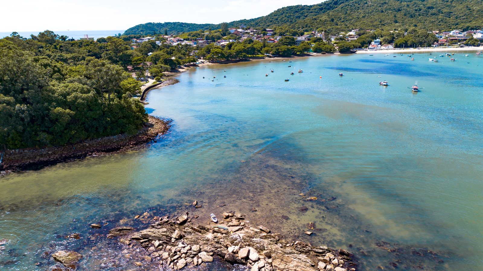Baía de água turquesa vista do alto, com costão de pedra em primeiro plano, barcos de pesca ancorados, faixa de areia ao fundo e morro coberto de mata com casas baixas espalhadas.