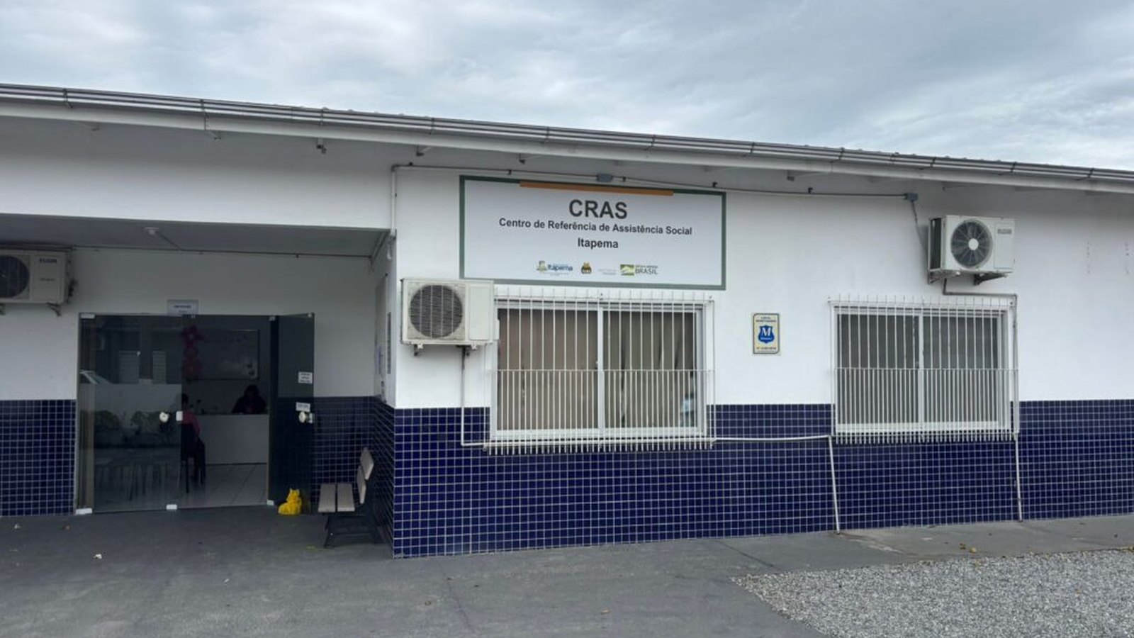 Fachada do Centro de Referência de Assistência Social de Itapema, prédio branco com faixa de azulejo azul, placa do CRAS acima da entrada e pátio de concreto à frente.