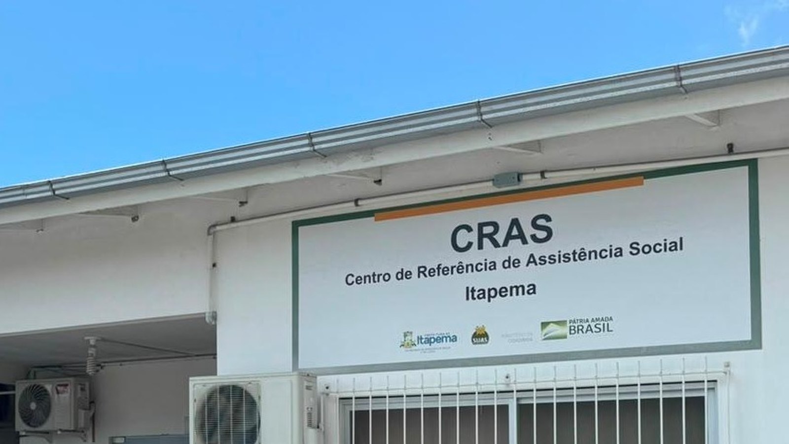 Fachada branca do CRAS de Itapema vista de baixo, com a placa do Centro de Referência de Assistência Social acima da entrada, telhado de calha aparente, aparelhos de ar-condicionado na parede e grade branca na janela, sob céu azul limpo.