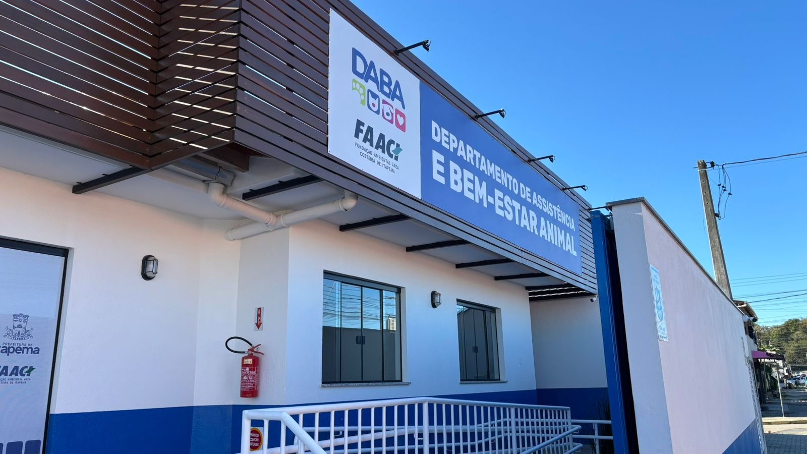 Fachada da sede do DABA em Itapema, pr&eacute;dio branco com faixa azul na parte de baixo, letreiro azul escrito Departamento de Assist&ecirc;ncia e Bem-Estar Animal sobre a entrada e uma rampa de corrim&atilde;o branco na frente, em dia de c&eacute;u limpo.
