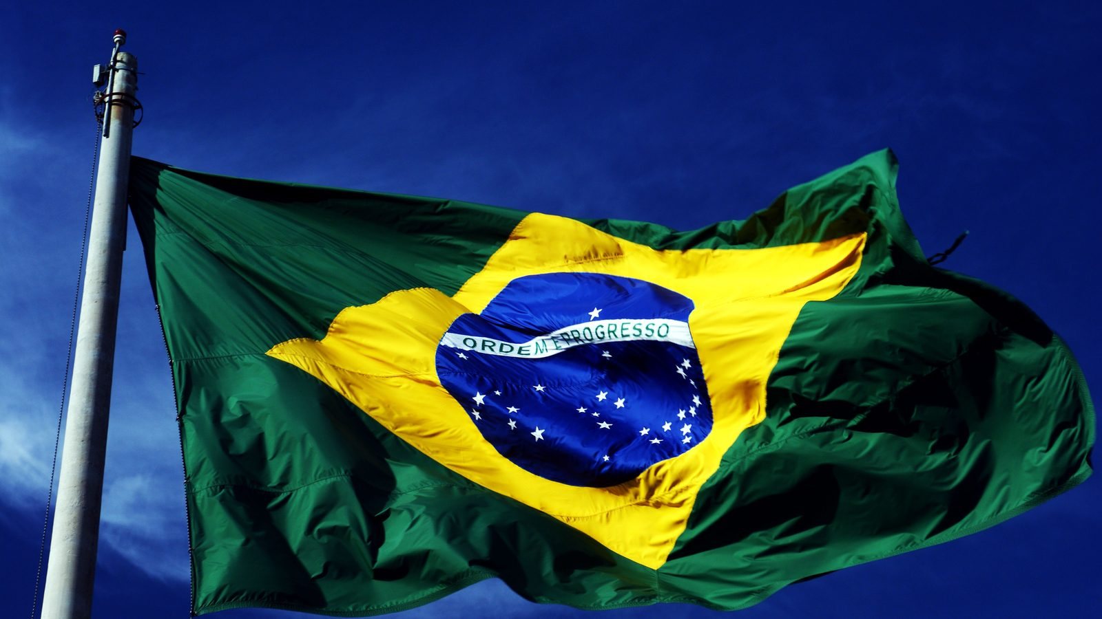 Bandeira do Brasil hasteada em mastro de metal, tremulando aberta pelo vento contra um céu azul sem nuvens.