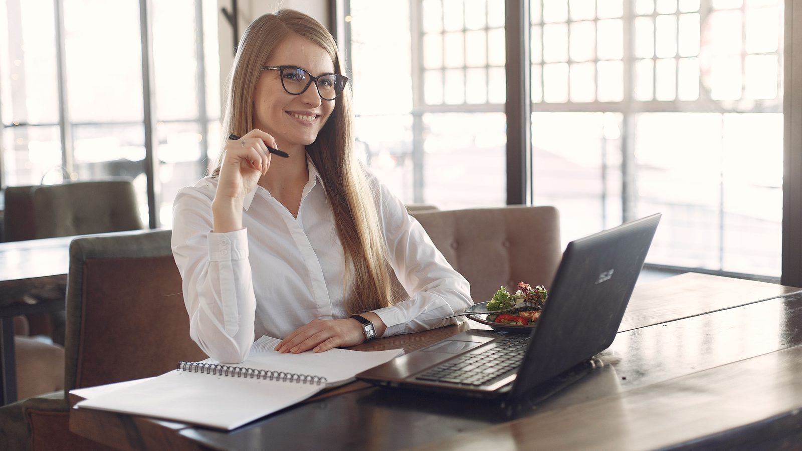 Mulher de camisa branca e óculos sentada a uma mesa de madeira, sorrindo com uma caneta na mão, ao lado de um caderno aberto e de um notebook ligado.