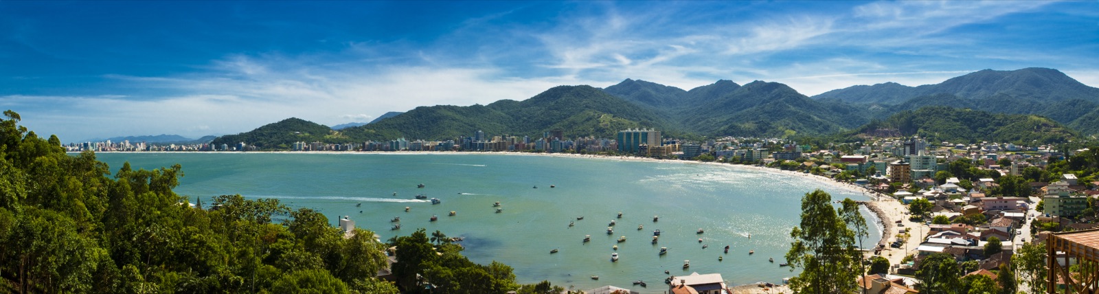 Vista panorâmica da enseada de Itapema a partir de um ponto alto, com o mar à esquerda, a faixa de areia ao fundo e morros cobertos de mata contornando a baía.