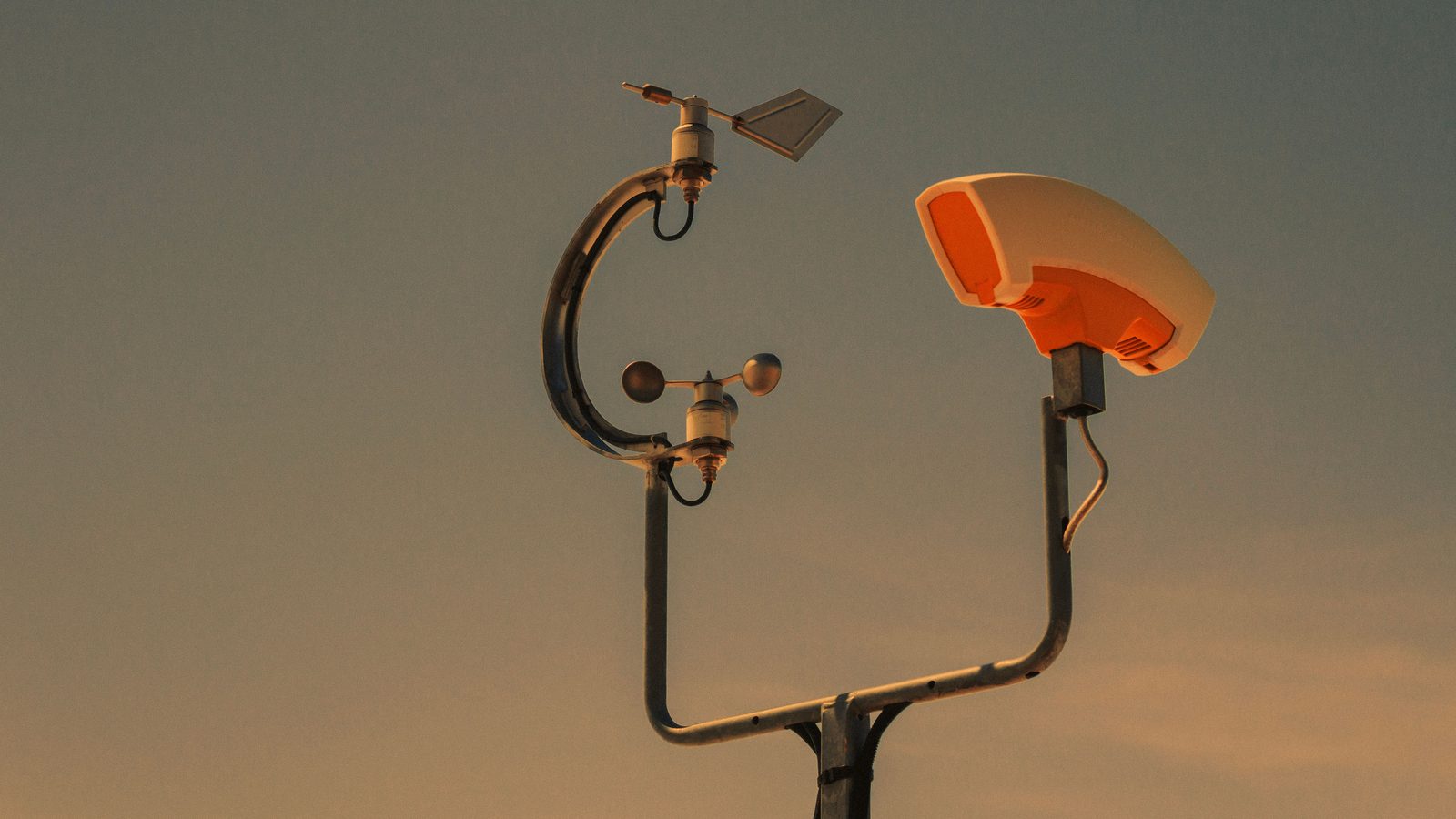 Topo de uma estação meteorológica recortada contra o céu no fim da tarde, com o cata-vento de direção e o anemômetro de três conchas presos a um braço curvo de metal e, ao lado, o abrigo branco do sensor de temperatura, tudo montado num mastro.