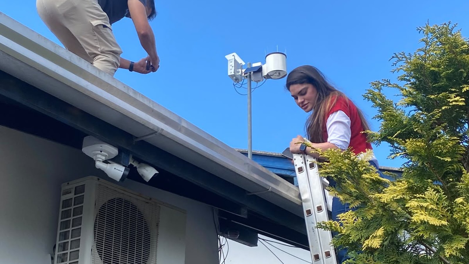 Dois técnicos instalam uma estação meteorológica no telhado de uma escola sob céu azul: um homem agachado sobre a calha aperta uma peça e uma mulher de colete vermelho trabalha no alto de uma escada de alumínio, com o mastro da estação, o pluviômetro branco e a placa solar ao fundo.