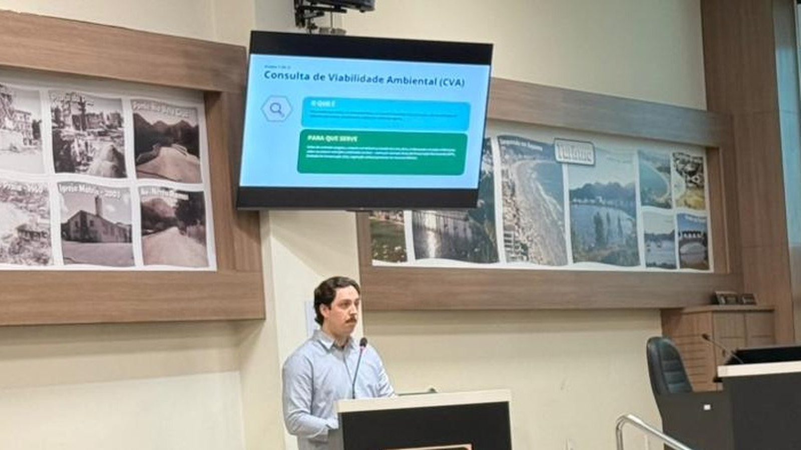 Técnico de camisa clara fala na tribuna do auditório da Câmara de Itapema, com um telão ao lado mostrando o slide Consulta de Viabilidade Ambiental (CVA) e painéis de fotos históricas da cidade na parede.