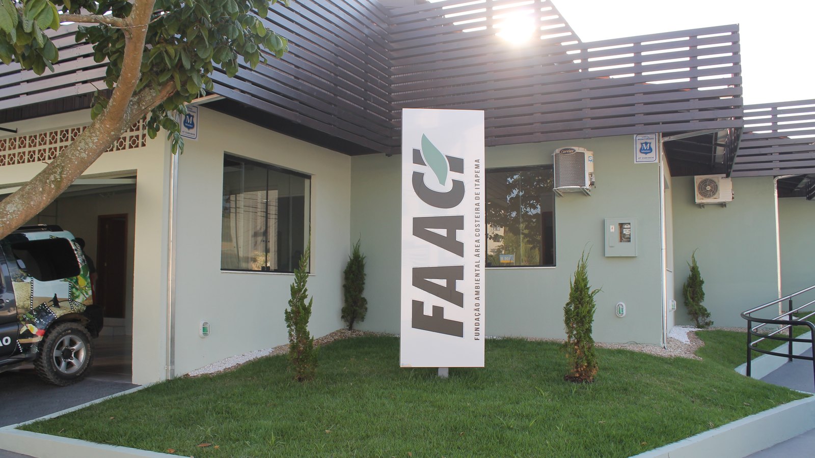 Fachada da sede da FAACI em Itapema, casa de paredes verde-claras com brise de madeira escura no telhado, gramado na frente e um totem branco com a sigla FAACI em letras grandes.