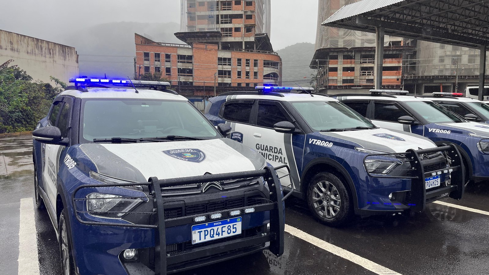 Cinco viaturas da Guarda Municipal de Itapema estacionadas em fila num pátio molhado de chuva, a da frente com giroflex azul aceso e grade de proteção preta na frente, prédios em obra e morro com neblina ao fundo.
