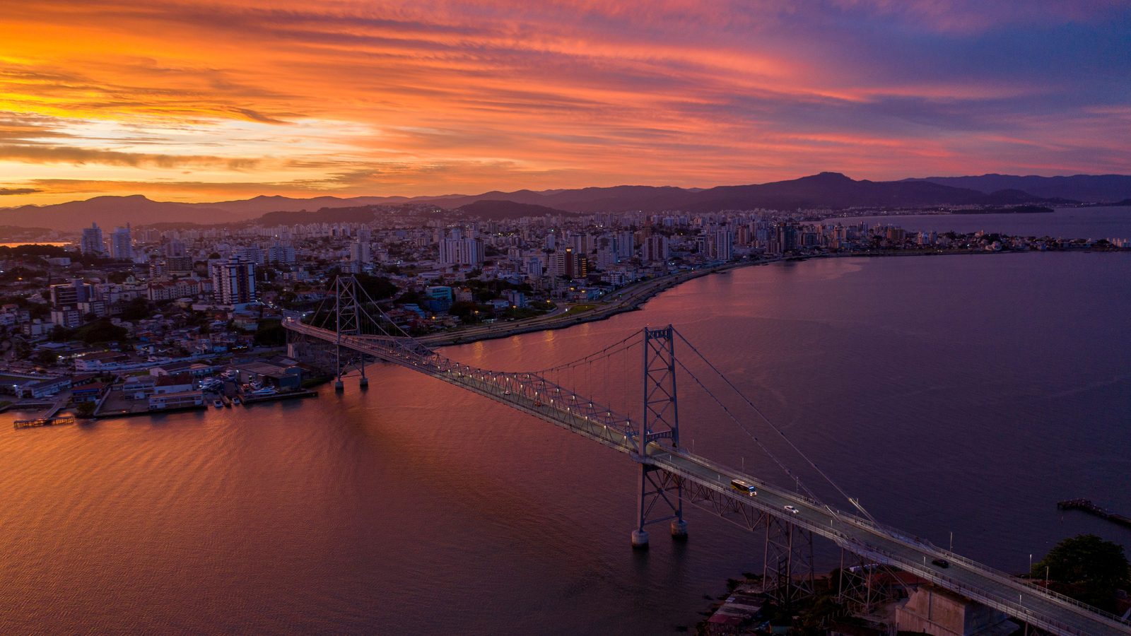 Ponte pênsil metálica iluminada ao pôr do sol, vista do alto, ligando duas margens sobre o mar calmo, com a cidade ao fundo.