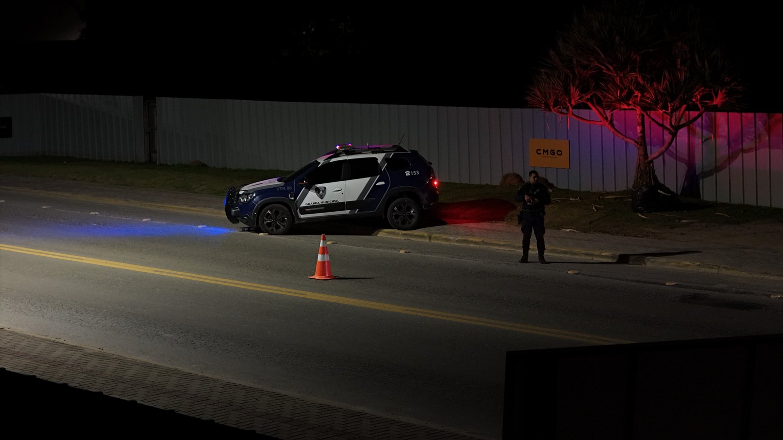 Viatura da Guarda Municipal de Porto Belo parada à beira da rua à noite, com giroflex azul e vermelho aceso, um cone laranja no asfalto e um agente fardado em pé na calçada ao lado.
