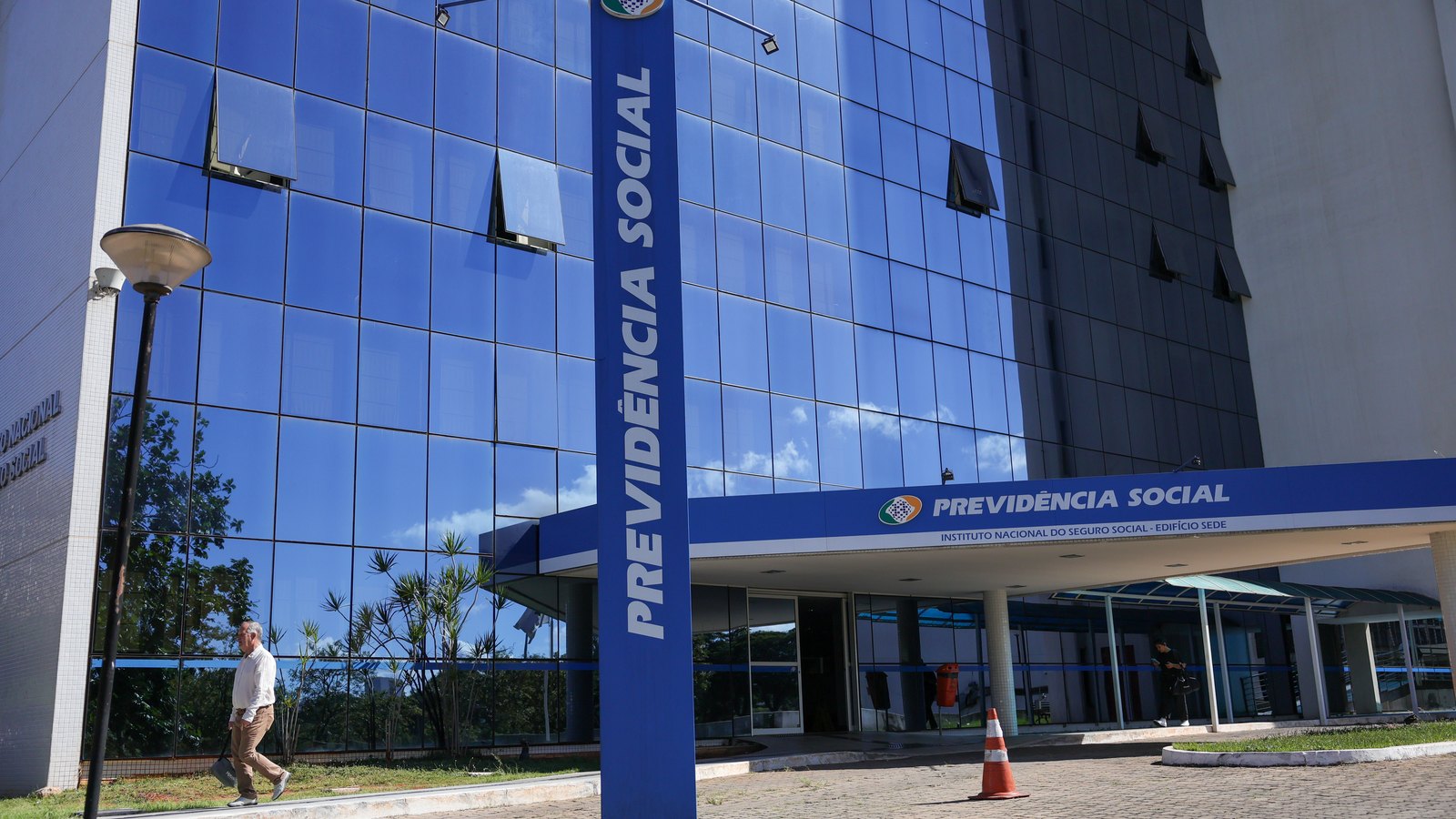 Fachada envidraçada do edifício sede do INSS em Brasília, com um totem azul alto escrito Previdência Social na frente e o céu refletido nos vidros.