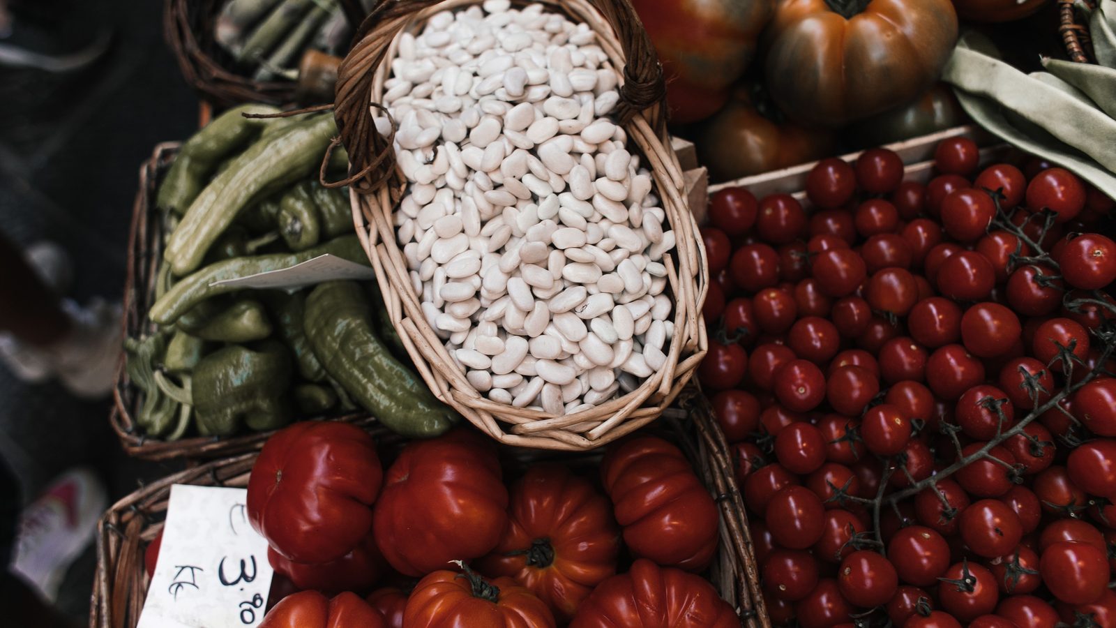 Banca de feira vista de cima, com caixas de tomate vermelho maduro, tomate-cereja no ramo, pimentões verdes e um cesto de feijão branco, com uma etiqueta de preço escrita à mão apoiada na frente.