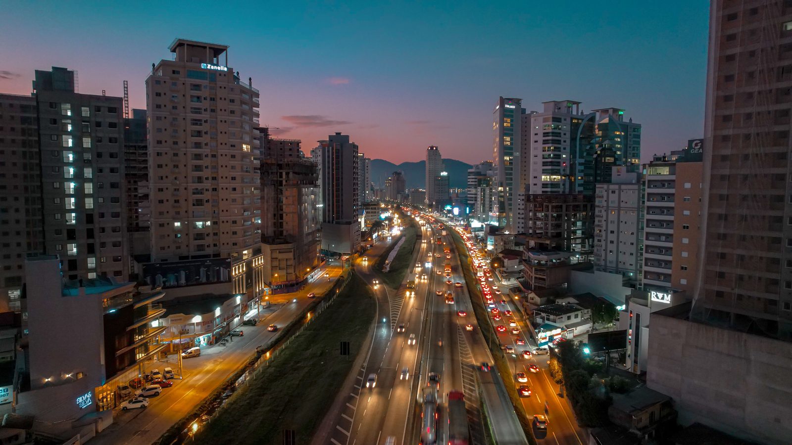 Itapema ao anoitecer vista do alto: a BR-101 corta a imagem ao meio com as duas pistas cheias de carros de faróis acesos, e prédios altos se erguem dos dois lados.
