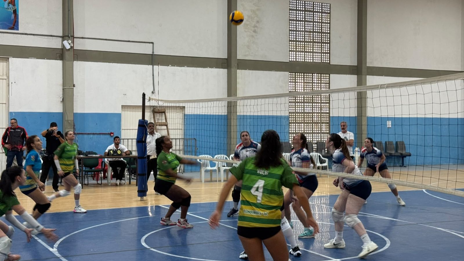 Partida de voleibol feminino em ginásio, com a bola no alto sobre a rede e jogadoras de duas equipes posicionadas em quadra azul.