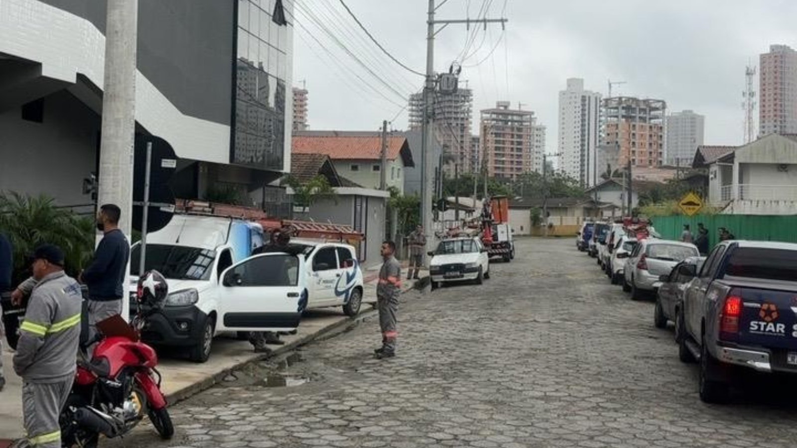 Rua de paralelepípedo em Porto Belo num dia nublado, com poste de energia carregando um feixe de cabos que cruza a via, carros estacionados dos dois lados, um caminhão com cesto aéreo ao fundo e trabalhadores de uniforme cinza parados na calçada.