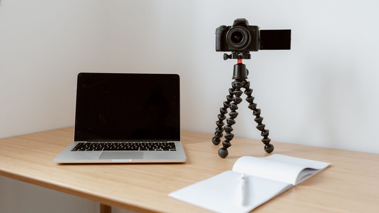 Mesa de madeira clara com um notebook aberto de tela apagada, uma câmera fotográfica montada num tripé flexível ao lado e um caderno branco aberto à frente, diante de uma parede lisa.