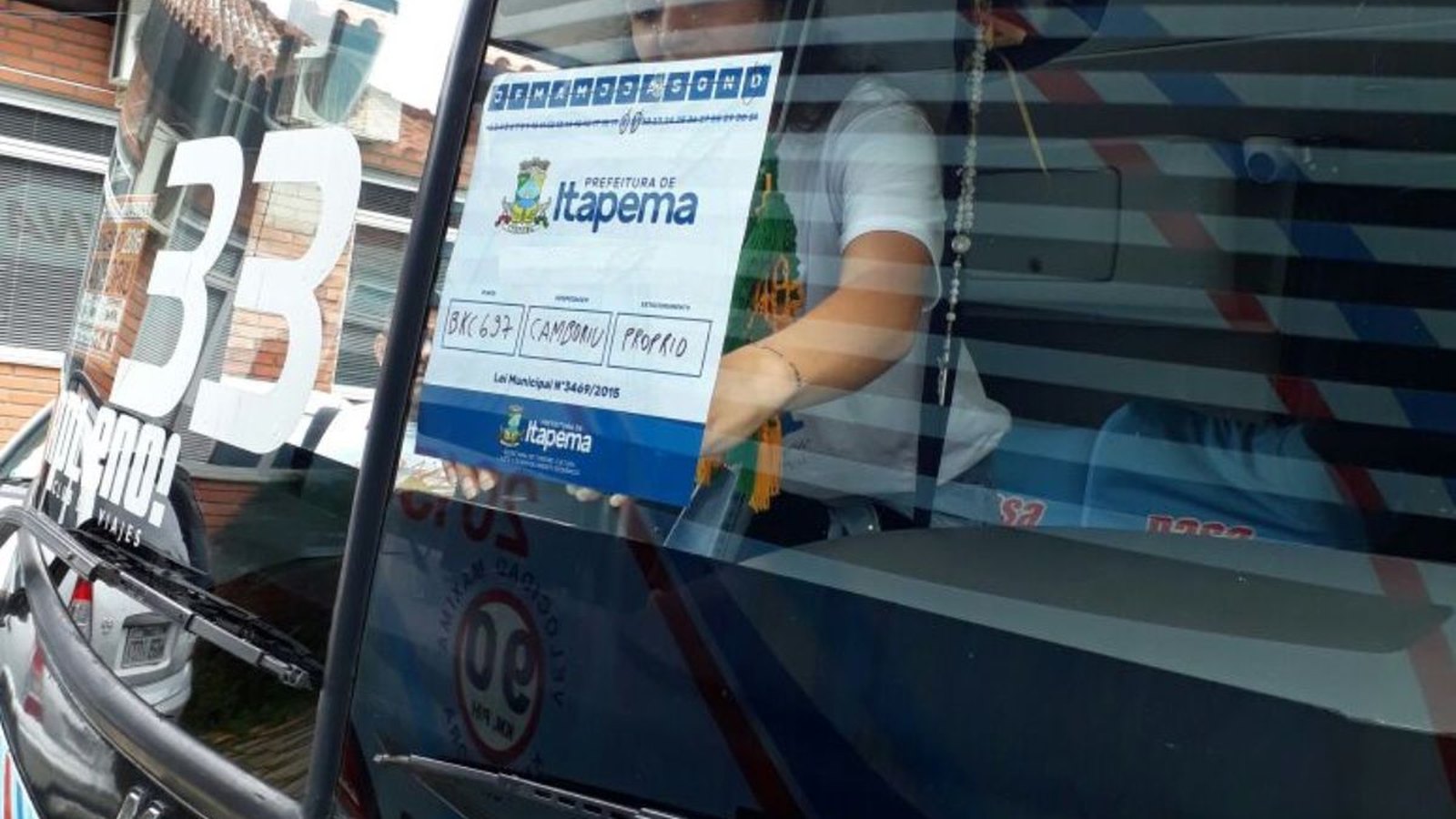 Selo de papel da Prefeitura de Itapema colado por dentro no para-brisa de um ônibus de excursão, com o brasão do município, campos preenchidos à mão com a placa e a cidade de origem do veículo e a citação da lei municipal no rodapé.