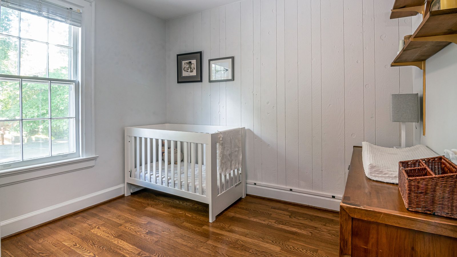 Quarto de bebê vazio, com berço branco de grades encostado na parede de madeira clara, colchão firme e plano coberto por lençol branco, janela à esquerda e cômoda de madeira escura à direita.