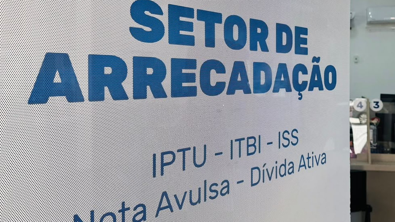 Placa adesivada no vidro do Setor de Arrecadação da Prefeitura de Itapema, com as palavras SETOR DE ARRECADAÇÃO em letras azuis grandes e, abaixo, a linha IPTU, ITBI, ISS, Nota Avulsa e Dívida Ativa.