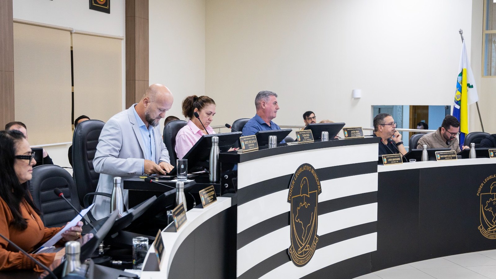 Vereadores de Itapema sentados na bancada curva e preta do plenário durante uma sessão, com placas de identificação douradas, microfones de haste, garrafas térmicas e monitores à frente de cada lugar, e a bandeira do município ao fundo.