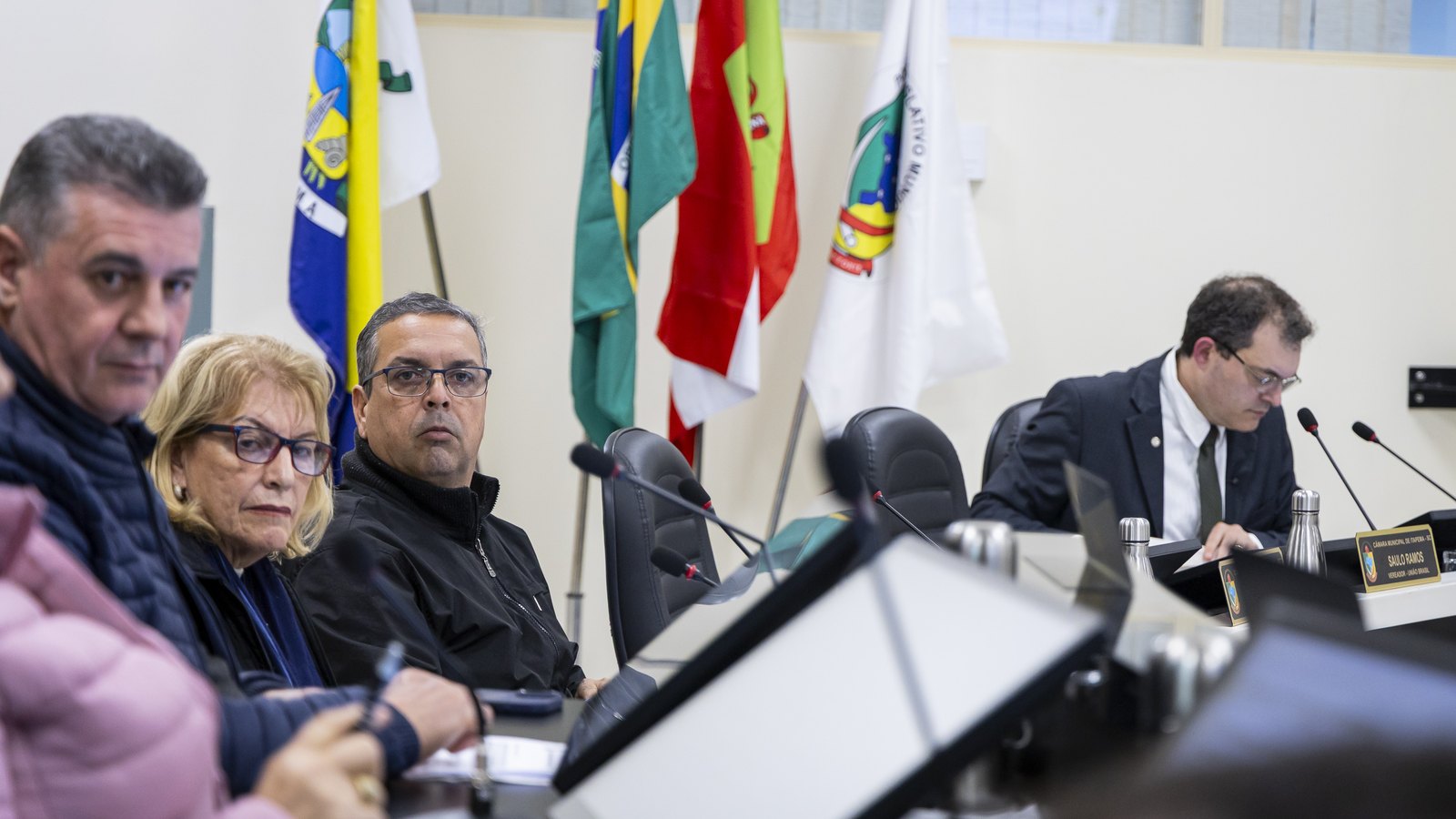 Vereadores de Itapema sentados lado a lado na bancada do plenário durante uma sessão, com microfones de haste, garrafas térmicas e monitores à frente, as bandeiras do Brasil, de Santa Catarina e do município ao fundo e uma placa de identificação dourada à direita.