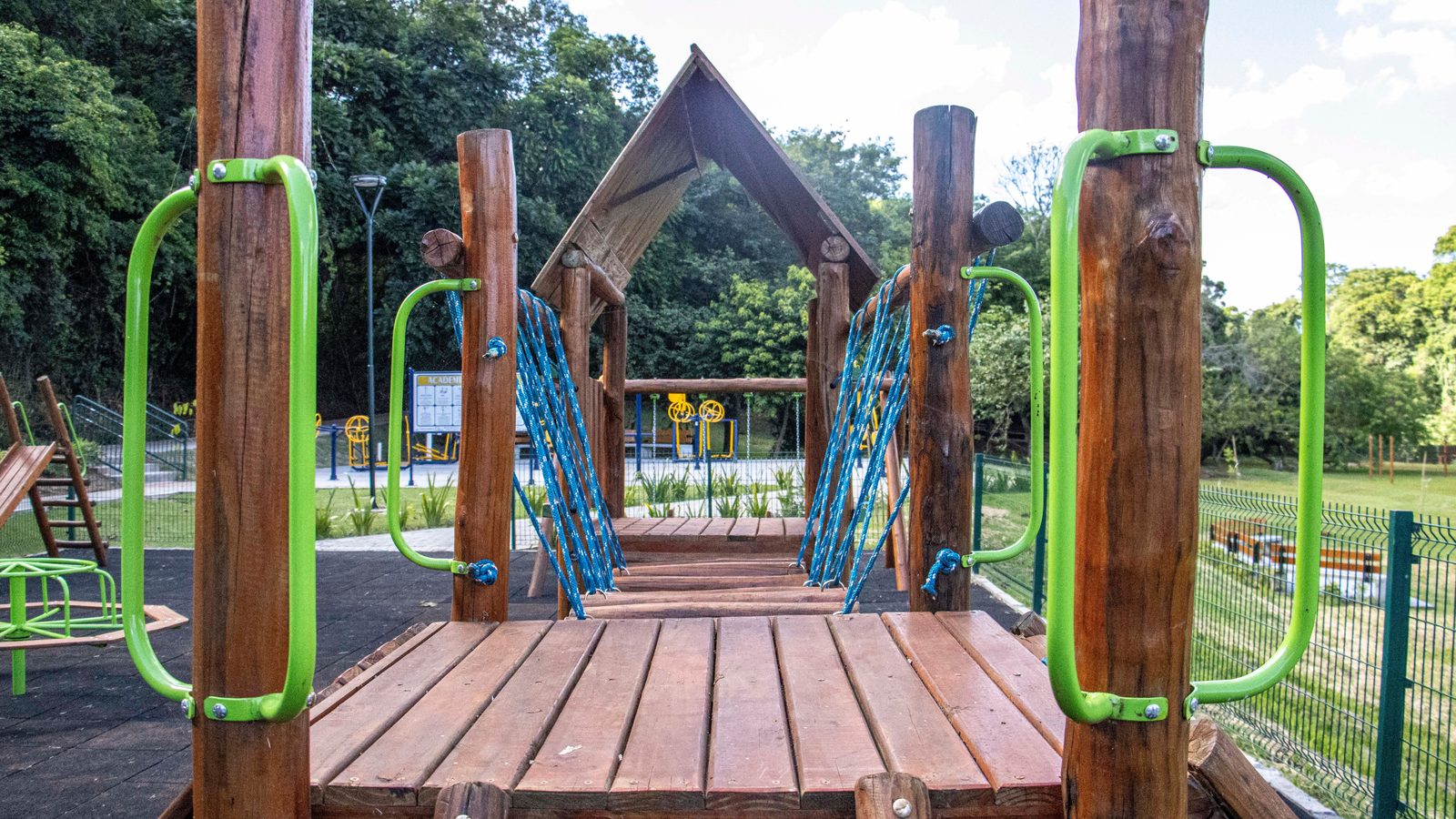 Estrutura de madeira de parquinho vista de frente, com plataforma de tábuas, postes de tronco roliço, corrimãos de metal verde e uma ponte de cordas azuis ao fundo, dentro de uma praça cercada por grama, árvores e aparelhos de academia ao ar livre, sem ninguém no quadro.