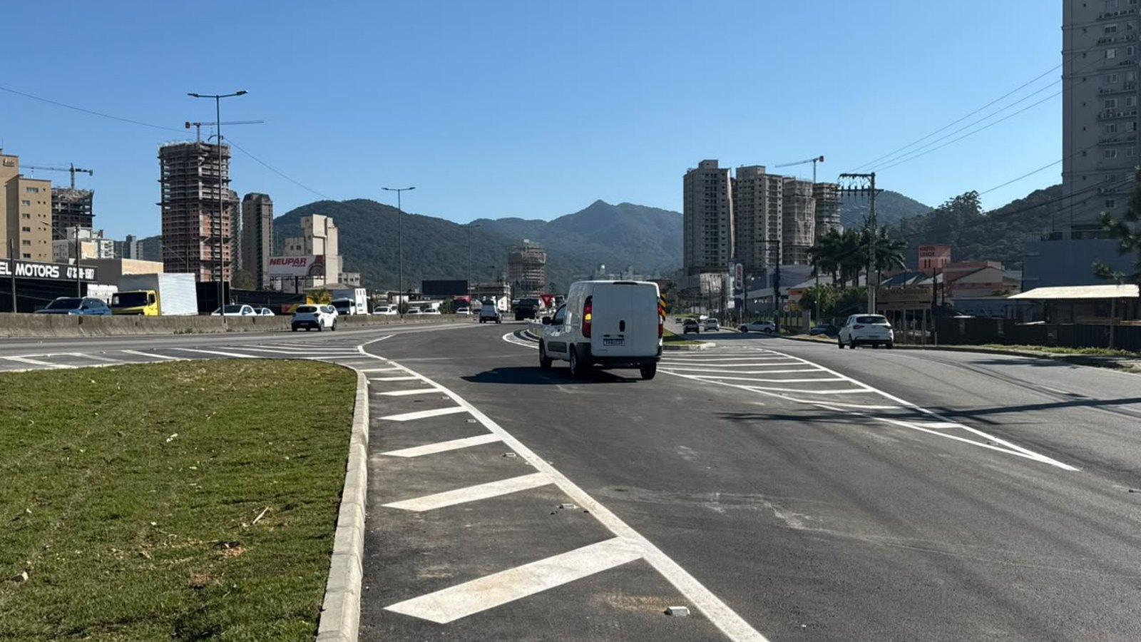 Alça nova de acesso à BR-101 em Itapema recém-aberta, com asfalto escuro e faixas brancas de zebrado pintadas no chão, uma van branca passando pela pista, meio-fio e gramado à esquerda, prédios altos e morros verdes no horizonte sob céu azul.