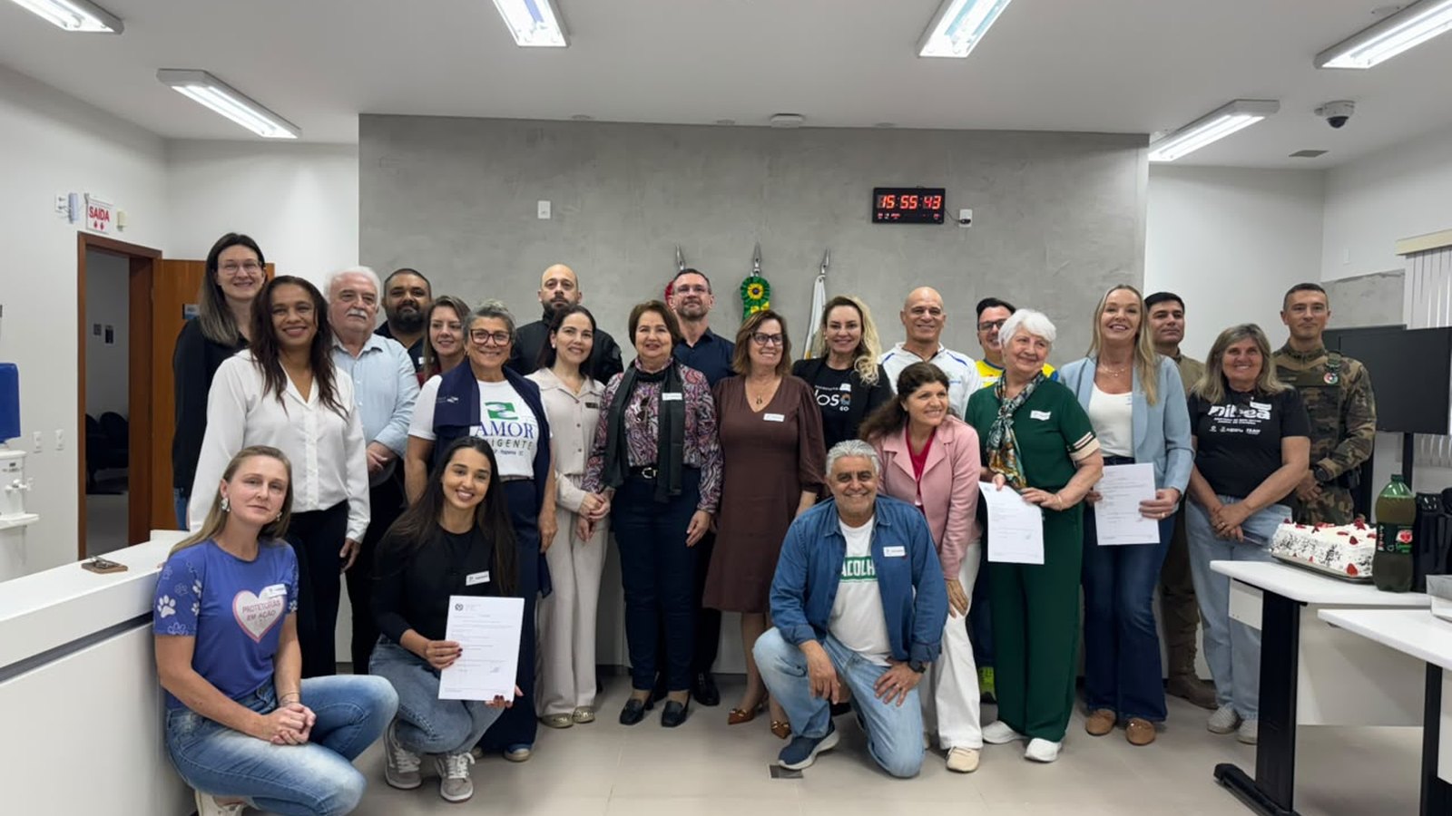 Cerca de vinte e cinco pessoas adultas posam para foto de grupo numa sala clara do Fórum de Itapema, algumas segurando folhas de papel brancas nas mãos, com bandeira do Brasil ao fundo, relógio digital vermelho na parede e luminárias de teto acesas.