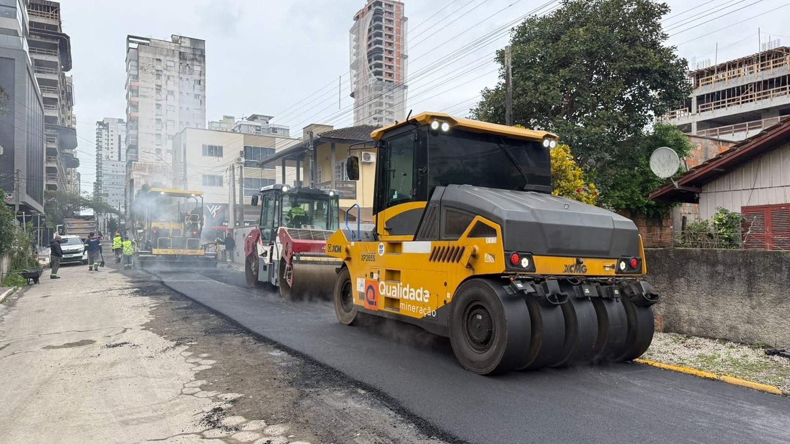 Rolo compactador amarelo passando sobre asfalto novo e escuro numa rua de Itapema, com uma pavimentadora e um caminhão mais adiante, fumaça branca subindo do asfalto quente, operários de colete refletivo ao fundo, prédios altos à esquerda e casas baixas à direita sob céu nublado.