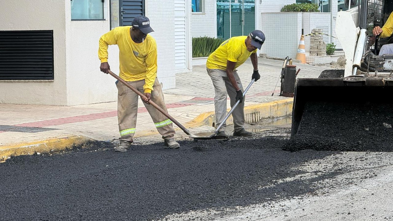 Dois trabalhadores de camiseta amarela e boné espalham massa asfáltica preta com rodos numa rua de Itapema, diante de um prédio residencial branco, enquanto a caçamba de uma pá-carregadeira despeja mais asfalto à direita e a mancha de asfalto novo cobre quase toda a largura da pista.