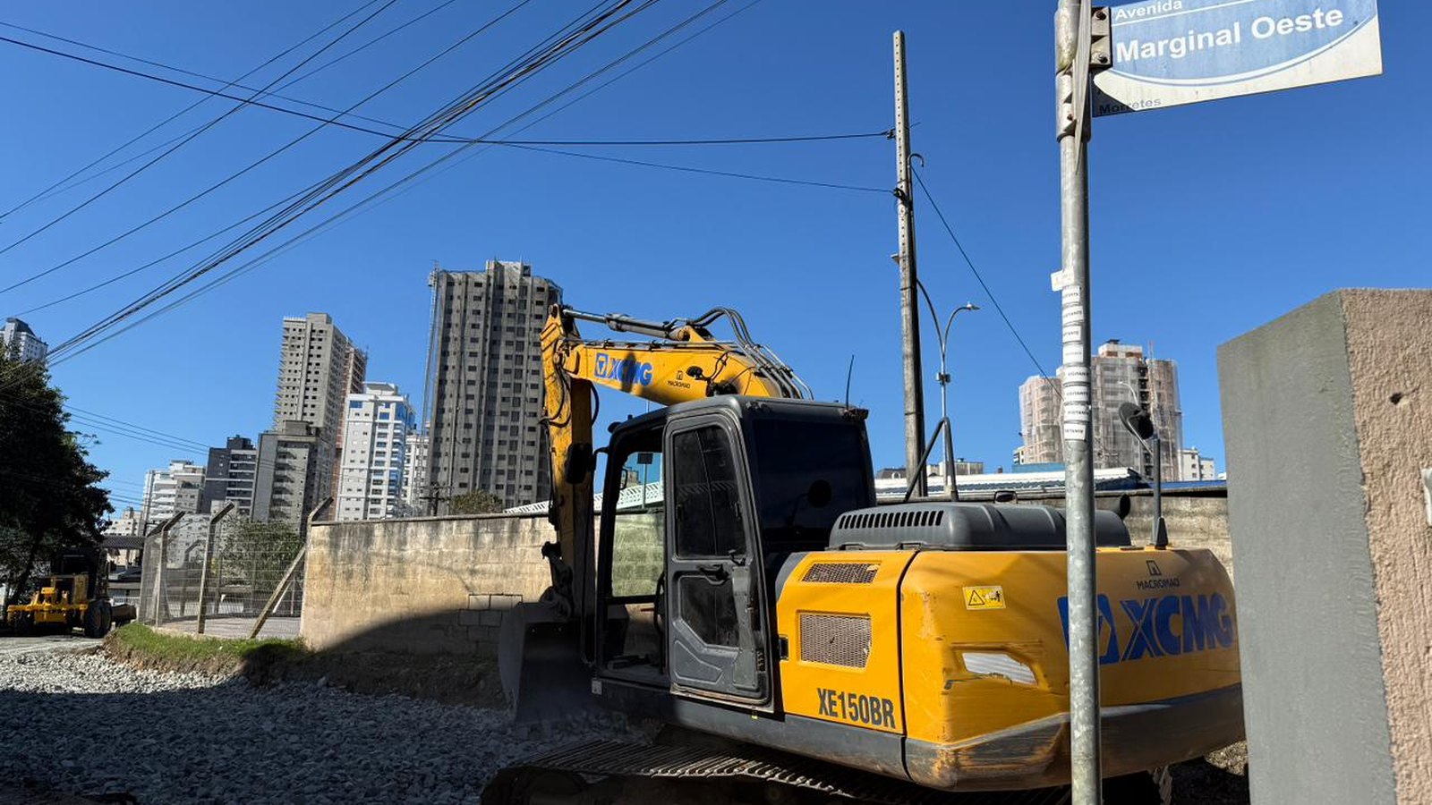 Escavadeira amarela parada sobre o leito de terra e brita da Avenida Marginal Oeste, no bairro Morretes, ao lado da placa azul de rua com o nome da avenida, com prédios altos ao fundo e céu azul limpo.